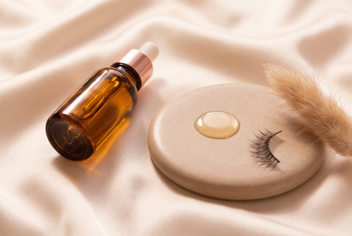 Huile de ricin dorée dans un flacon compte-gouttes ambré, goutte d'huile et cils naturels sur fond crème — soin des cils Lashéa